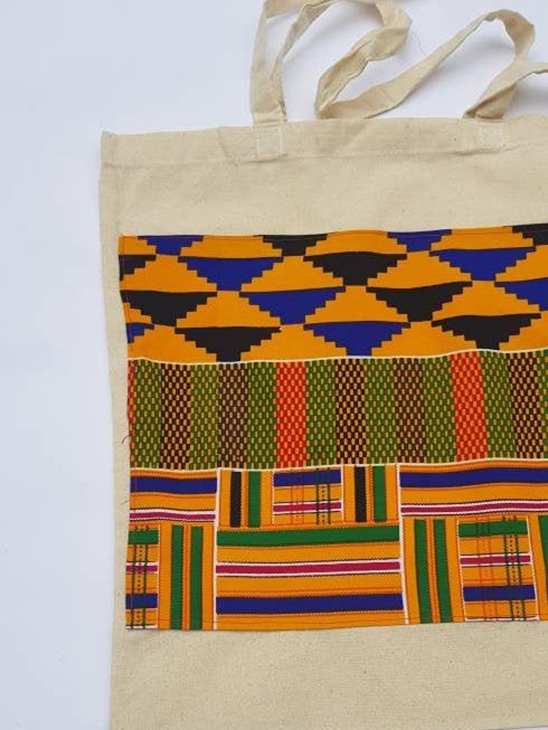 May include: A natural canvas tote bag with a large pocket on the front. The pocket is made of colorful African print fabric with geometric patterns in shades of orange, green, blue, and black.