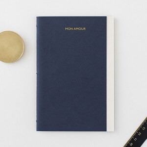 Könnte beinhalten: Ein dunkelblaues Notizbuch mit weißem Rücken und dem Schriftzug "MON AMOUR" in goldenen Lettern auf dem Einband.
