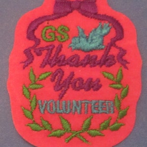 May include: A red felt patch with embroidered text and design. The patch features a purple bow, the letters "GS" in green, a teal bird, and the words "Thank You VOLUNTEER" in purple and teal. A green leaf wreath surrounds the word "VOLUNTEER".