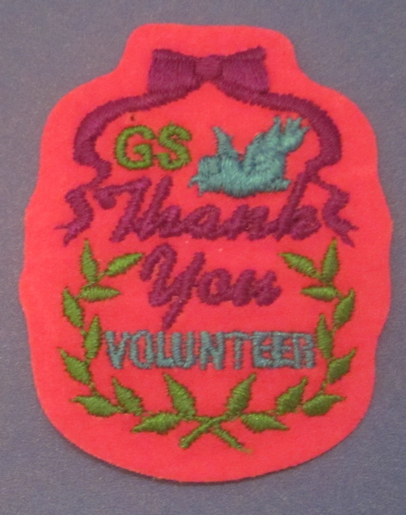 May include: A red felt patch with embroidered text and design. The patch features a purple bow, the letters "GS" in green, a teal bird, and the words "Thank You VOLUNTEER" in purple and teal. A green leaf wreath surrounds the word "VOLUNTEER".