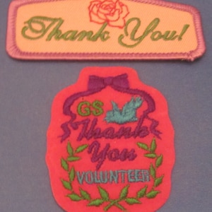 May include: Two embroidered patches. The top patch is rectangular with a pink border, peach center, and green text that says "Thank You!" with a rose graphic. The bottom patch is red with purple accents, a blue bird, and the words "Thank You Volunteer".