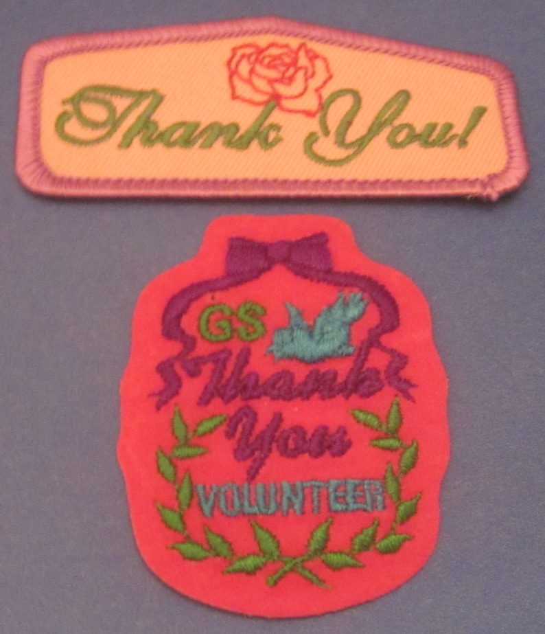 May include: Two embroidered patches. The top patch is rectangular with a pink border, peach center, and green text that says "Thank You!" with a rose graphic. The bottom patch is red with purple accents, a blue bird, and the words "Thank You Volunteer".