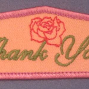 May include: Embroidered patch with a peach-colored background and a pink border. The patch features the words "Thank You!" in green script, and a red rose illustration.