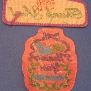 May include: Two embroidered patches. The top patch is rectangular with a purple border, a flower, and the word "Birthday" in green. The bottom patch is a pink, bottle-shaped design with "Happy Birthday" text.