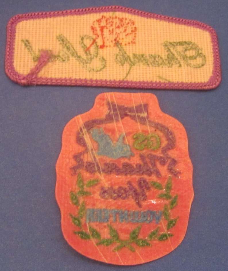 May include: Two embroidered patches. The top patch is rectangular with a purple border, a flower, and the word "Birthday" in green. The bottom patch is a pink, bottle-shaped design with "Happy Birthday" text.