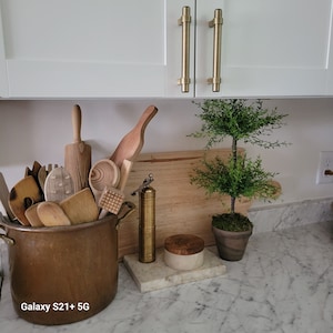 May include: A kitchen scene featuring a copper pot filled with wooden utensils, a rolling pin, cutting board, and a small potted topiary. A brass pepper grinder and a marble salt cellar are also present.