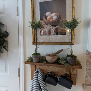 May include: A rustic kitchen display featuring a framed picture of eggs in a nest, a wooden shelf with a mortar and pestle, two small potted trees, and two black mugs. A striped towel hangs below.