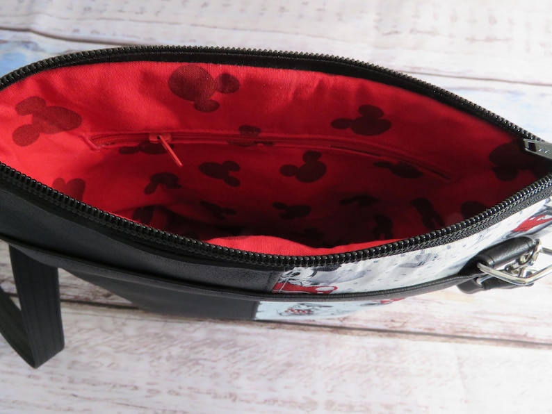 Puede incluir: Bolso bandolera negro con forro rojo y blanco con estampado de Mickey Mouse. El bolso tiene cierre de cremallera y una correa.
