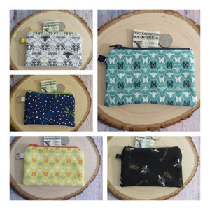 May include: Four small zippered pouches with different fabric patterns. Each pouch has a zipper closure and a small loop for attaching a key ring. The pouches are filled with cash and coins. The patterns include bees, butterflies, and dragonflies.