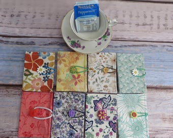 May include: Eight rectangular fabric pouches with various floral and botanical patterns, each secured with a colorful elastic band and a decorative button. Above the pouches sits a white teacup containing a Bigelow Peppermint herbal tea bag.