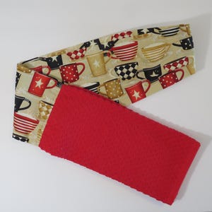 May include: A kitchen towel with a red, textured bottom and a patterned top. The top fabric features various coffee mugs in red, black, and gold on a beige background. The towel is folded in half.