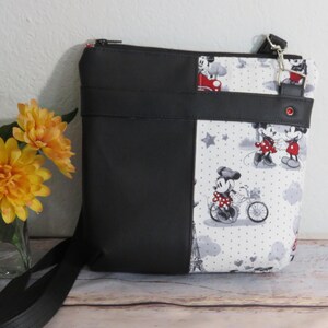 Puede incluir: Bolso bandolera negro y blanco con estampado de Mickey Mouse y Minnie Mouse. El bolso tiene una cremallera negra y una correa negra.