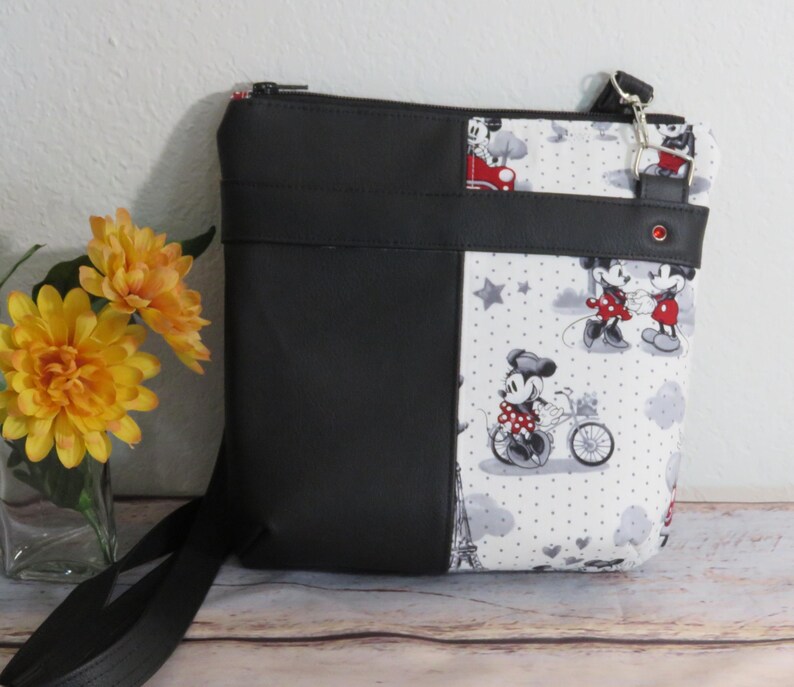 Puede incluir: Bolso bandolera negro y blanco con estampado de Mickey Mouse y Minnie Mouse. El bolso tiene una cremallera negra y una correa negra.