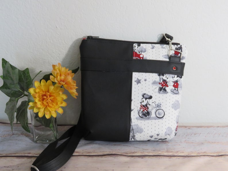 Puede incluir: Bolso bandolera de Mickey Mouse y Minnie Mouse en blanco y negro con cremallera y correa negras. El bolso tiene un panel de tela negra en la parte delantera.