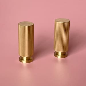 Puede incluir: Dos pomos de armario de metal dorado con un diseño acanalado. Los pomos tienen una base redonda y una forma cilíndrica.