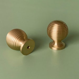 May include: Two gold-toned cabinet knobs with a ribbed design. The knobs are round and have a smooth, polished finish.