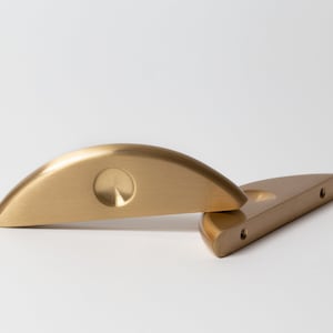 Puede incluir: Dos tiradores de armario de metal dorado con forma redondeada de medio círculo. El tirador superior tiene un diseño circular hundido en el centro. El tirador inferior tiene dos agujeros para tornillos.