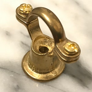 May include: A brass-coloured metal eye bolt with a rounded loop and two screws. The base is circular, and the bolt is designed to be screwed into a surface. The hardware is shown against a marble background.