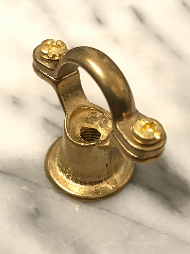 May include: A brass-coloured metal eye bolt with a rounded loop and two screws. The base is circular, and the bolt is designed to be screwed into a surface. The hardware is shown against a marble background.