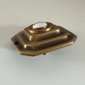 May include: A vintage brass doorbell button with a white ceramic center. The button has a stepped, geometric design and a tarnished finish, suggesting age. The doorbell is likely for a home's front door.