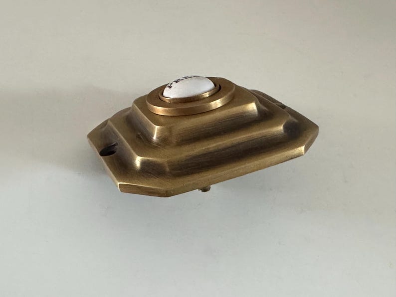May include: A vintage brass doorbell button with a white ceramic center. The button has a stepped, geometric design and a tarnished finish, suggesting age. The doorbell is likely for a home's front door.