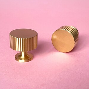 May include: Two gold-colored cabinet knobs with a ribbed design on a pink background. One knob is upright, and the other is lying on its side. The knobs have a cylindrical shape with a flat top and a short, rounded base.