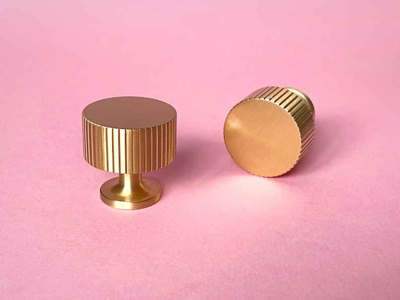 May include: Two gold-colored cabinet knobs with a ribbed design on a pink background. One knob is upright, and the other is lying on its side. The knobs have a cylindrical shape with a flat top and a short, rounded base.