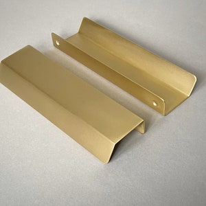 May include: Two gold metal cabinet pulls with a rectangular shape and rounded edges. The pulls are made of a brushed metal and have a matte finish.