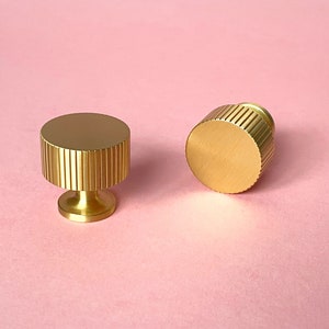 May include: Two gold-colored cabinet knobs with a fluted design. One knob is upright, and the other is on its side. The knobs have a cylindrical shape with a flat top and a small, round base. The background is a solid pink color.