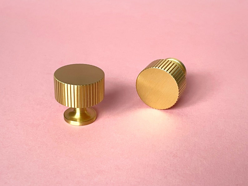 May include: Two gold-colored cabinet knobs with a fluted design. One knob is upright, and the other is on its side. The knobs have a cylindrical shape with a flat top and a small, round base. The background is a solid pink color.