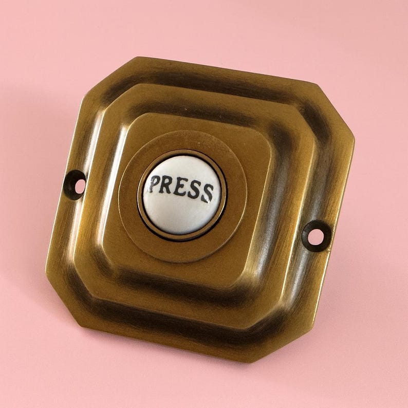 May include: A vintage-style doorbell button with a bronze finish. The button has a white, circular center with the word "PRESS" in black lettering. The doorbell is square with rounded edges and two screw holes.