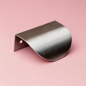 Puede incluir: Un tirador de armario de metal cepillado con un diseño curvo. El tirador es plateado y tiene un pequeño agujero en el lateral. El fondo es de color rosa claro liso.