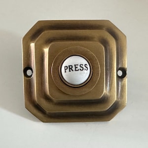 May include: A vintage brass doorbell button with a square, stepped design. The center features a white button with the word "PRESS" in black. Two screw holes are visible on either side of the button.