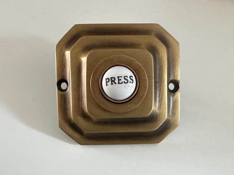 May include: A vintage brass doorbell button with a square, stepped design. The center features a white button with the word "PRESS" in black. Two screw holes are visible on either side of the button.