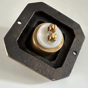 May include: A close-up of a brass and white object with two screws, set within a dark, geometric frame. The object has a cylindrical shape with a white top and brass base. The frame is dark and has a geometric shape.
