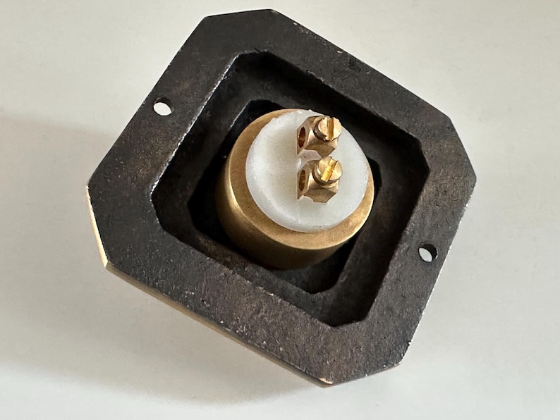 May include: A close-up of a brass and white object with two screws, set within a dark, geometric frame. The object has a cylindrical shape with a white top and brass base. The frame is dark and has a geometric shape.