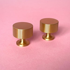 May include: Two gold-colored cabinet knobs with a cylindrical, ridged design. The knobs have a flat, circular top and a short, rounded base. They are set against a soft pink background, highlighting their metallic finish.