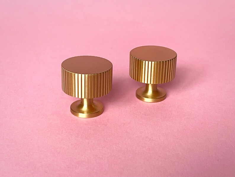 May include: Two gold-colored cabinet knobs with a cylindrical, ridged design. The knobs have a flat, circular top and a short, rounded base. They are set against a soft pink background, highlighting their metallic finish.