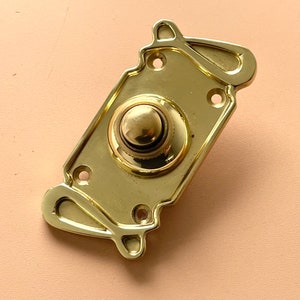 Peut inclure: Un bouton de sonnette en or avec un bouton rond et surélevé au centre. La sonnette a un design décoratif et orné.