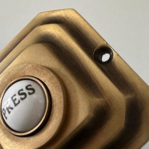 May include: Close-up of a vintage brass doorbell button. The button has a white center with the word "PRESS" in black lettering. The surrounding plate is a geometric design with a dark, aged finish.