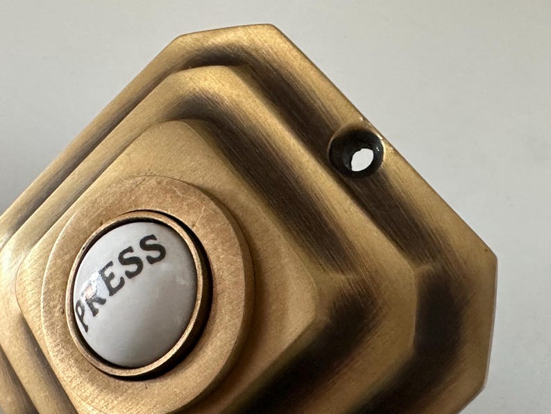 May include: Close-up of a vintage brass doorbell button. The button has a white center with the word "PRESS" in black lettering. The surrounding plate is a geometric design with a dark, aged finish.