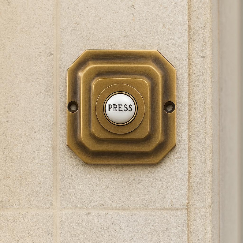 May include: A square, antique brass doorbell button with a white center. The center button is round and has the word "PRESS" printed on it. The doorbell is mounted on a light beige tiled wall.