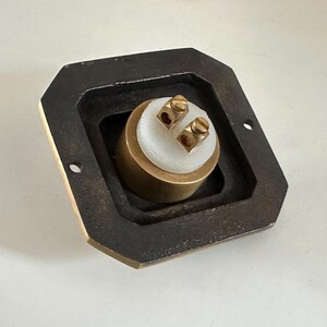 May include: A brass electrical component with two screw terminals, set within a dark, rectangular frame. The component has a white central section and is designed for electrical connections. The frame has two mounting holes.