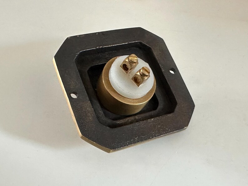 May include: A brass electrical component with two screw terminals, set within a dark, rectangular frame. The component has a white central section and is designed for electrical connections. The frame has two mounting holes.