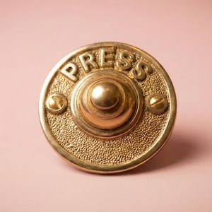 May include: A vintage brass doorbell with the word "PRESS" embossed on the front. The doorbell has a textured surface and is secured with two screws. The button is in the center, and the overall design is circular.