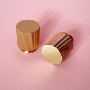 May include: Two gold-colored cabinet knobs with a ribbed design. The knobs are round and have a smooth, polished finish.