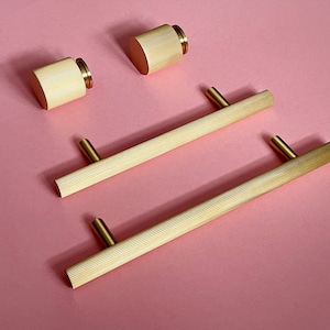 Könnte beinhalten: Vier goldfarbene Schrankbeschläge, darunter zwei lange, gerippte Schubladenzüge und zwei runde, gerippte Knöpfe. Die Beschläge werden auf einer rosa Oberfläche präsentiert.