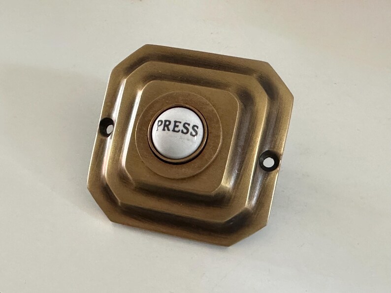May include: Antique brass doorbell button with a white center. The button is inscribed with the word "PRESS" in black lettering. The doorbell has a square shape with rounded edges and two screw holes.