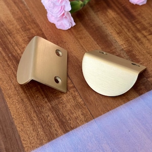 May include: Two brass-colored corner brackets with a rounded design. Each bracket has two pre-drilled holes for mounting. The brackets are made of metal and have a matte finish. The background is a wooden surface.