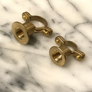 May include: Two brass pipe clamps with a threaded interior and a circular ring. The clamps are made of a shiny, golden metal and are set against a white and grey marble background.